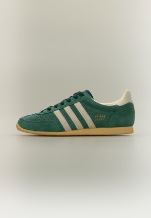 Grøn ruskind Adidas Japan sneaker med hvide striber, beige hæl og gummisål vist fra siden på en ensfarvet baggrund.