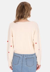 Maglioncino beige chiaro lavorato a maglia con ricamo di cuori rossi sulle maniche. Presenta una vestibilità rilassata, scollo ampio e orlo a coste. Abbinato a una gonna di jeans blu.