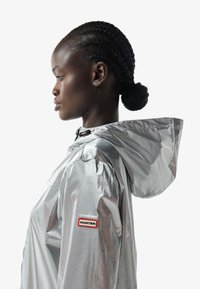 Perfil de una persona con cabello trenzado que lleva un impermeable plateado Hunter con capucha y parche con el logo en la manga, sobre un fondo liso.