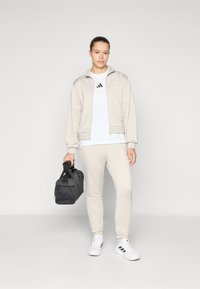 Lys beige treningsjakke med matchende joggebukser, hvit t-skjorte med svart logo, og svart bæreveske. Hvite sneakers fullfører antrekket.