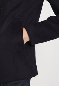 Abrigo de lana azul marino con un bolsillo lateral, cuello redondeado y detalle de botón negro en el dobladillo, con una textura suave y un corte entallado.