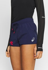 Pantalones cortos deportivos azul marino con cintura de cordón, que presentan un logo rojo en el lado izquierdo y un logo plateado en el derecho. Tejido ligero.