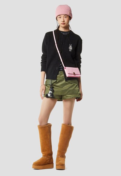 Νέα γυναίκα που φοράει ροζ σκούφο, μαύρη ζακέτα με πατς, χακί σορτς, καφέ γούνινες μπότες μέχρι το γόνατο και ροζ τσάντα χιαστί.