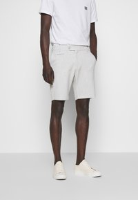 Male model wearing beige tailored shorts, white t-shirt, and white sneakers, standing against a plain white background.