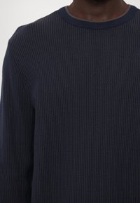 Homme portant un pull en maille texturée bleu marine foncé à encolure ronde, sur un fond clair uni.