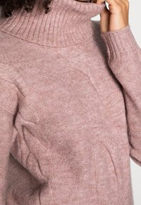 Pull en tricot rose avec un col roulé côtelé haut, présentant des motifs en relief sur les côtés et un tissu doux et confortable.