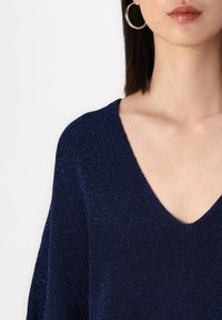 Marineblå stickad tröja med V-ringning och glittrande textur. Har vida ärmar och en avslappnad passform som framhäver en subtil glans.