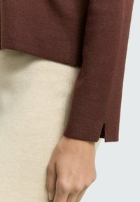 Pull en tricot marron avec une fente à l'ourlet, associé à une jupe beige clair. Le tissu est doux et texturé, offrant un aspect en couches.