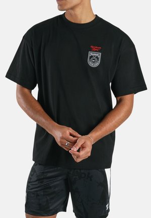 Hombre con camiseta negra de Reebok con logo rojo y blanco en el pecho y pantalones cortos negros con patrón, manos entrelazadas relajadamente al frente.