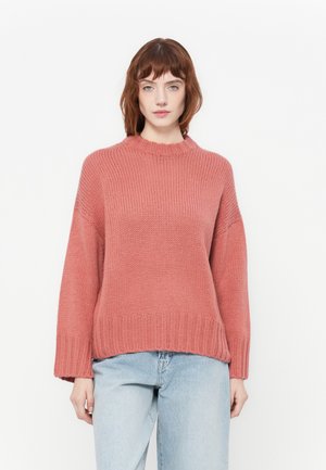 Teksturert rosa strikkegenser med høy hals og vide ermer, kombinert med lyseblå jeans. Bunnpartiet har ribbestrikket kant.