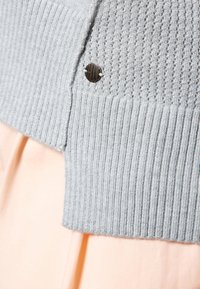 Close-up van een lichtgrijze gebreide cardigan met een kleine ronde zwart-gouden knoop op een licht perzikkleurige stof achtergrond.