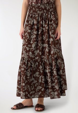 Gonna lunga marrone con stampa paisley bianca, vita arricciata, abbinata a sandali bassi marroni su piedi nudi.