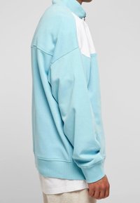 Ljusblå lös jacka med vit panel, hög krage och långa ärmar. Tillverkad av mjukt material med ribbade ärmslut och nederkant.