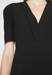 Robe noire avec un décolleté en V et des manches courtes bouffantes portée par une personne à la peau claire, vue des épaules à la taille.