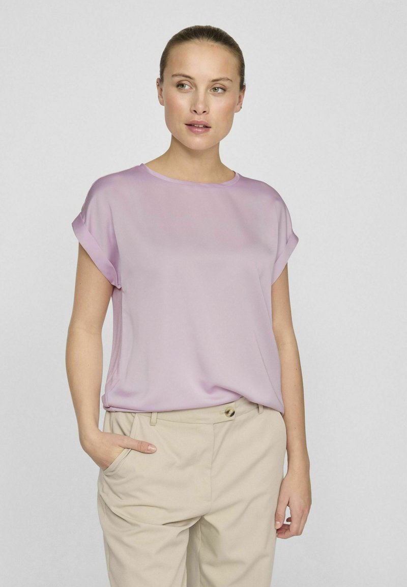 Blusa corta a maniche corte di colore viola chiaro con scollo rotondo, realizzata in tessuto morbido, abbinata a pantaloni beige con tasche laterali.