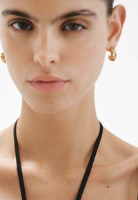 Pendientes de aro de oro con una superficie texturizada, que presentan un corte en forma de corazón. La modelo lleva una blusa negra de tirantes sencilla. Se aprecia una textura de piel suave.