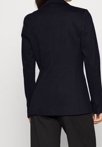 Blazer navy ajustado con tejido suave, hombros estructurados y cierre de un solo botón. Presenta un corte slim y mangas de longitud completa.