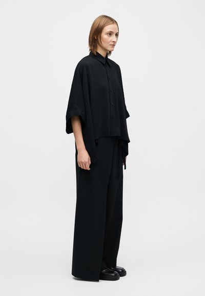 Chemise noire surdimensionnée avec col et boutons, associée à un pantalon noir à jambes larges. Les deux ont une texture lisse et un design minimaliste.