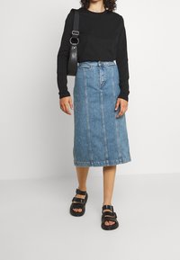 Jeanskjol i ljusblått, knälång med sömdetaljer, kombinerad med en svart långärmad skjorta och svarta sandaler. Enkelt design.