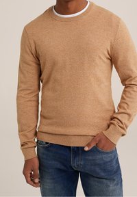 Brun texturerad tröja med en rundad halsringning och ribbade ärmslut, i kombination med blå denimjeans. Tröjan har en melerad tygfinish.