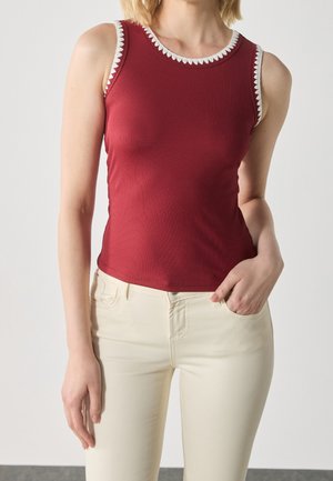 Kobieta w czerwonym, bezrękawnym prążkowanym topie z białym obszyciem i kremowych spodniach, stojąca z jedną ręką w kieszeni na jednolitym tle.