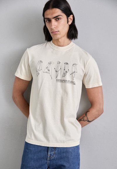 Cream cotton T-shirt featuring a black line-drawing graphic of figures in various poses, with "KIDSUPER STUDIO" text at the bottom.