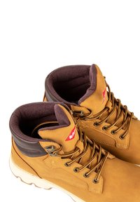 Scarponi da trekking in pelle marrone con colletto imbottito di colore marrone scuro, lacci color sabbia e suola in gomma bianca. Presenta un piccolo logo rosso sulla parte superiore.