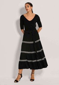 Schwarzes Kleid mit enganliegendem Oberteil, V-Ausschnitt und Ärmeln bis zum Ellbogen. Präsentiert einen gestuften Rock mit weißem gewelltem Saum und einem Taillengürtel.