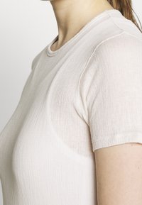 Světle béžové tričko s krátkým rukávem vyrobené z texturované, ribstované tkaniny. Má kulatý výstřih a přiléhavý střih.