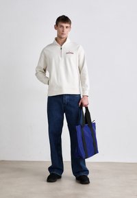 Sudadera crema con cremallera y texto bordado en rojo, jeans de mezclilla azul y una bolsa tote azul con asas negras. El modelo está de pie sobre un fondo claro.