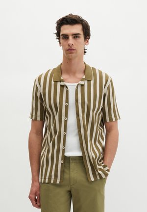 Giovane uomo che indossa una camicia a manica corta a righe verticali verdi e bianche sopra una canottiera bianca e pantaloni verde oliva, in piedi con la mano in tasca.