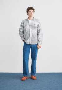 Καρό πουκάμισο με κουμπιά σε γκρι, λευκό και μπλε, συνδυασμένο με μπλε τζιν και πορτοκαλί μοκασίνια. Το μοντέλο στέκεται σε μπλε χαλί μπροστά σε λευκό φόντο.