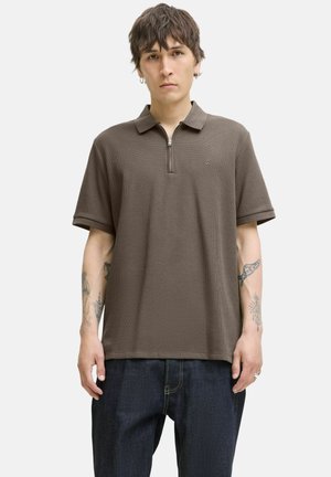 Jeune homme aux cheveux courts portant une chemise marron à manches courtes avec col zippé et un jean foncé, debout devant un fond clair uni, avec des tatouages sur les bras.