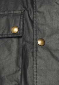 Detalj av svart vaxat tygjacka med en bröstficka och två mässingstryckknappar ingraverade med "Belstaff England."