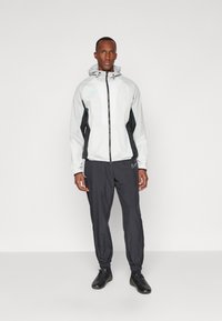 Veste légère Nike en gris clair avec des accents noirs, dotée d'une fermeture zippée, d'une capuche et de poignets élastiques. Assortie à un pantalon de jogging noir.