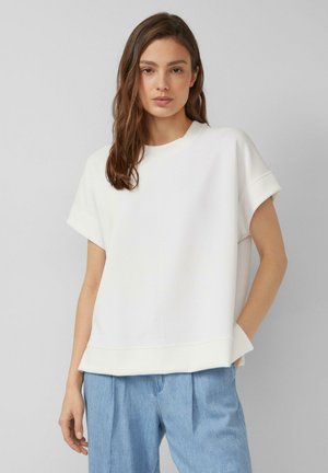 Femme aux longs cheveux bruns portant un haut blanc ample à manches courtes et un jean taille haute bleu clair, debout devant un fond neutre.