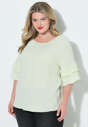 Frau mit welligem blondem Haar trägt eine hellgrüne Bluse mit Rüschenärmeln und schwarze Hose, posiert vor einem weißen Hintergrund.