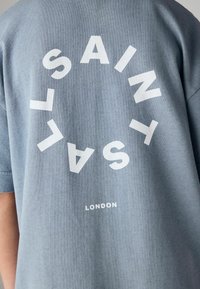 Chemise bleue avec des lettres blanches formant "SAINT SALV" en motif circulaire et "LONDON" imprimé en dessous au dos.