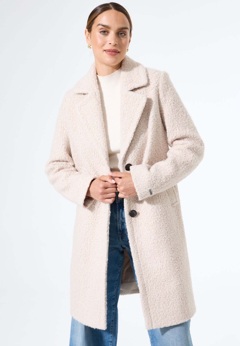 Licht beige, gestructureerde jas met een gelapte kraag, sluiting met knopen aan de voorkant en twee zijzakken, gedragen over een crèmekleurige top en blauwe jeans.