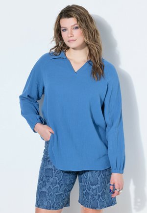 Vrouw met golvend haar draagt een losse blauwe blouse met lange mouwen en blauwe shorts met slangenprint, staand tegen een witte muur met hand in de zak.