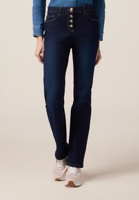 Jeans de tiro alto azul oscuro con cierre de cinco botones, piernas rectas y costuras en contraste, combinados con zapatillas claras sobre un fondo claro.