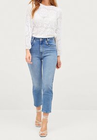 Eine Frau trägt ein weißes, langärmliges Lochspitzenoberteil, hochgeschnittene, hellblaue Jeans und weiße, schnüren Sandaletten mit Absatz. Sie steht vor einem schlichten Hintergrund.