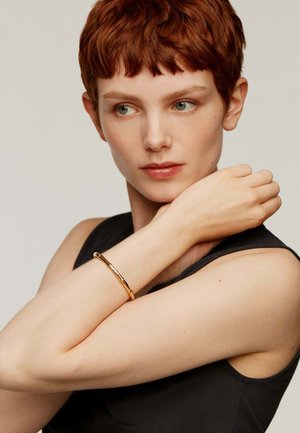 Brazalete de oro sobre un brazo pálido, con un diseño suave y minimalista, adornado con acentos ligeramente texturizados, exhibido sobre una blusa sin mangas oscura.