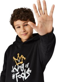 Svart hoodie med grafisk design som har en gyllene krona och texten "King of Fools" i vitt. Mjuk tyg, avslappnad passform och framficka.