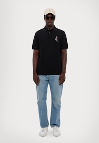 Cămașă polo neagră cu design brodat, blugi drepți ușor colorați în albastru deschis, adidași albi și o șapcă bej, completate de ochelari de soare negri.