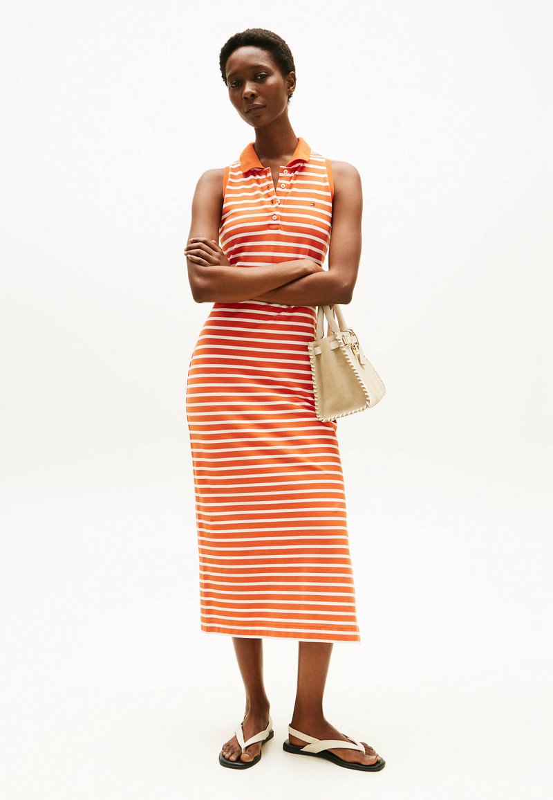 Sukienka bez rękawów w paski w kolorze pomarańczowym i białym, z kołnierzykiem i guzikami. Do tego lekka beżowa torebka i płaskie sandały.
