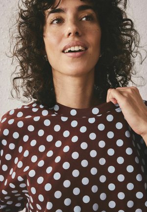 Tânără cu păr creț, purtând o cămașă maro cu buline albe mari, ținând ușor gulerul și privind ușor în sus.