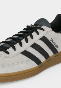 Άσπρο sneaker Adidas Spezial με μαύρες ρίγες, μαύρα κορδόνια και ανοιχτό καφέ λαστιχένια σόλα, εμφανίζεται από την πλευρά και από εμπρός.