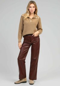 Femme debout, mains dans les poches, portant un pull en maille camel avec des boutons et un pantalon en cuir marron, associé à des bottines beiges sur fond gris.