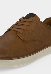 Gros plan d'une chaussure à lacets en cuir marron avec une couture détaillée et une semelle en caoutchouc blanche sur un fond clair.